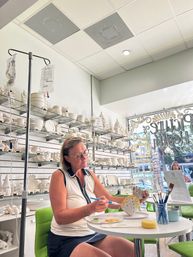 Woman painting a ceramic pineapple at a small table in a bright pottery-painting studio, shelves of white bisque ceramics behind her, paintbrushes, palette and green chairs by a sunny storefront window.