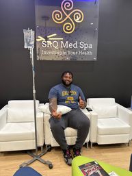 Person relaxed in a med spa lounge receiving IV infusion therapy, giving a thumbs-up while seated on a white armchair under a modern wellness sign, with white seating and wood floor visible.