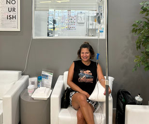 Smiling woman in a casual tank top receiving an IV infusion in a modern hydration therapy lounge with white chairs, side table with drink and takeout, gray walls and plant.