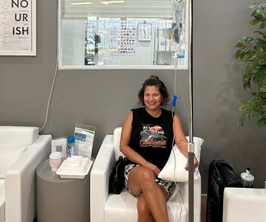Smiling woman in a casual tank top receiving an IV infusion in a modern hydration therapy lounge with white chairs, side table with drink and takeout, gray walls and plant.