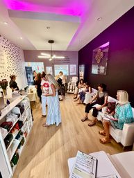 Bright med-spa waiting room with women seated and mingling, purple accent wall and pink LED ceiling, retail skincare shelves and massage chairs