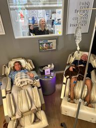 Two adults relaxing in beige massage chairs at a wellness lounge; one connected to an IV drip, both smiling under cozy blankets while a staff member waves through a service window.