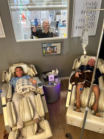 Two adults relaxing in beige massage chairs at a wellness lounge; one connected to an IV drip, both smiling under cozy blankets while a staff member waves through a service window.