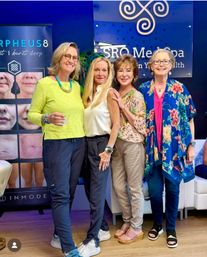 Four cheerful women pose together at a medspa reception, wearing colorful casual outfits and sandals with a blue branded wall and treatment posters visible behind them.