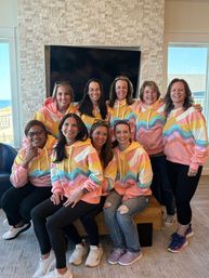 Nine women in matching pastel rainbow wave hoodies smiling for a group photo in a bright coastal living room with a stone fireplace, wall-mounted TV and ocean view through the windows.