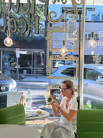 Artist painting a ceramic mug inside a bright downtown storefront studio with a large decorated window, hanging Edison bulbs, green chairs and a sidewalk view with parked cars.