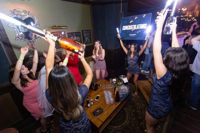 Women at a lively nightclub celebrating a bachelorette-style party with bottle service and sparklers, a congratulations sign held up, and drinks on a VIP table.