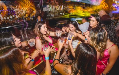 Group of women in party dresses toasting with shot glasses and a champagne flute at a crowded bar, colorful motion-blur lights and beer taps visible behind the counter, lively nightlife celebration.