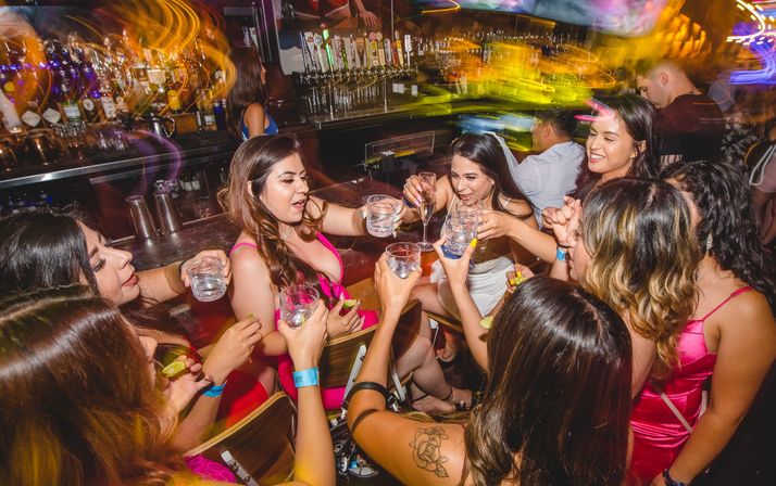 Group of women in party dresses toasting with shot glasses and a champagne flute at a crowded bar, colorful motion-blur lights and beer taps visible behind the counter, lively nightlife celebration.