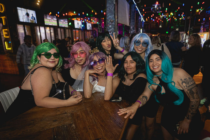 Bachelorette party at a neon-lit bar — seven friends smiling around a wooden table, bride-to-be with veil and headband, colorful wigs and sunglasses, lively nightlife atmosphere