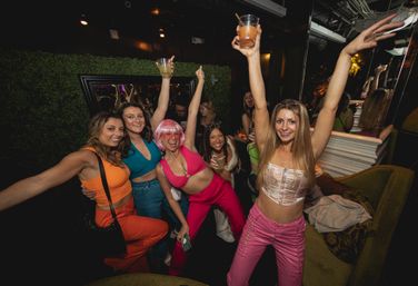 Group of friends in colorful outfits partying with drinks and raised arms in a lively nightclub lounge — fun girls' night-out city nightlife vibe