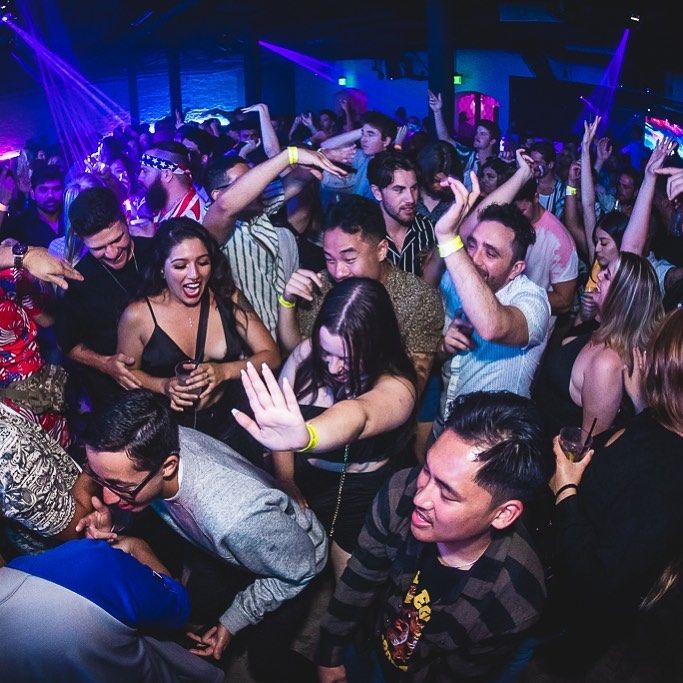 Packed nightclub dance floor with a crowd dancing and cheering under purple and blue neon lights, wristbands and drinks visible in a lively city nightlife scene.