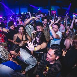 Packed nightclub dance floor with a crowd dancing and cheering under purple and blue neon lights, wristbands and drinks visible in a lively city nightlife scene.