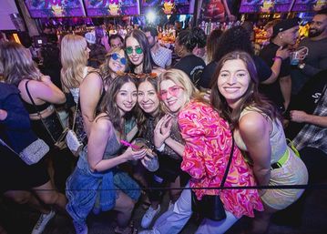Smiling group of friends in colorful outfits posing with drinks at a crowded neon-lit nightclub bar, lively urban nightlife party scene.
