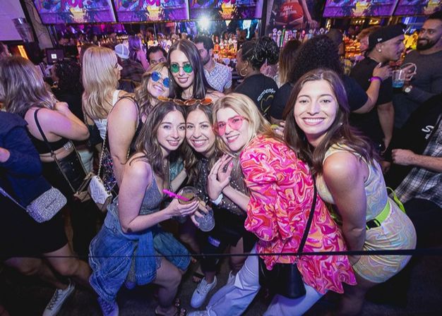 Smiling group of friends in colorful outfits posing with drinks at a crowded neon-lit nightclub bar, lively urban nightlife party scene.