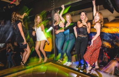 Six women dancing on a raised platform in a crowded nightclub with colorful motion-blur lights and a lively party atmosphere