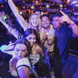Energetic group of young adults smiling and dancing at a vibrant nightclub bar under purple-blue lights, holding drinks and posing on a crowded dance floor.