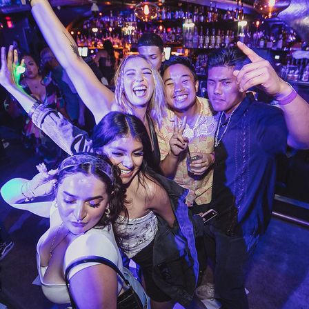 Energetic group of young adults smiling and dancing at a vibrant nightclub bar under purple-blue lights, holding drinks and posing on a crowded dance floor.
