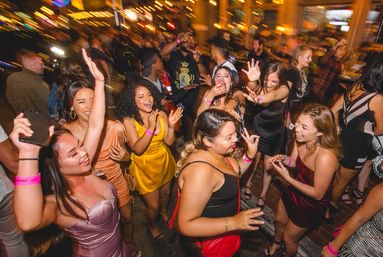 City nightlife dance party with a group of young women in colorful dresses laughing and dancing under warm string lights in a crowded outdoor bar scene