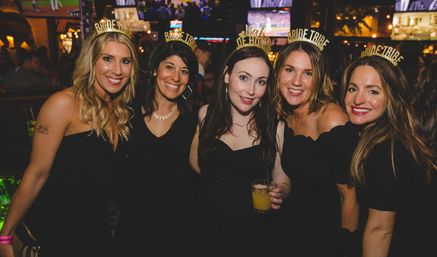 Five friends in black dresses wearing gold "Bride Tribe" and "Bride" headbands, smiling and holding cocktails at a lively bar bachelorette party.