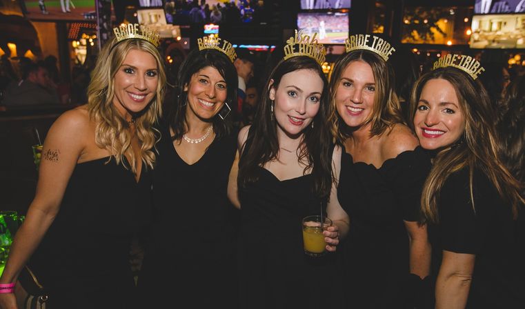 Five friends in black dresses wearing gold "Bride Tribe" and "Bride" headbands, smiling and holding cocktails at a lively bar bachelorette party.