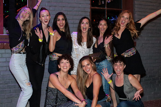 Nine women cheering outside a brick-walled bar at night, wearing casual party outfits and holographic fanny packs, one wearing a bride-to-be sash—energetic bachelorette night out.