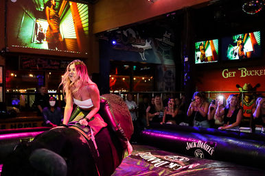 Woman laughing while riding a mechanical bull in a crowded bar as onlookers cheer and take photos under colorful neon lights and video screens.