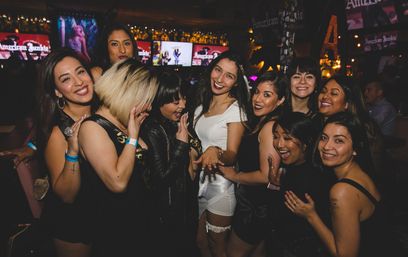 Smiling group of women at a lively nightclub bar celebrating a bachelorette, bride-to-be in a white dress and garter showing her ring while friends cheer.