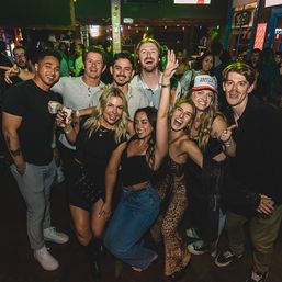 Group of friends cheering and posing with drinks at a crowded nightclub bar — lively nightlife scene.