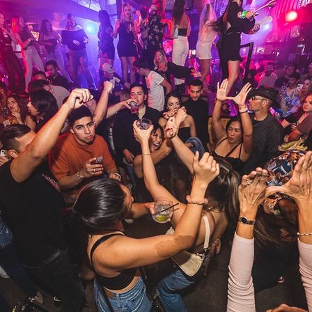 Packed urban nightclub dance floor under purple-pink lights, crowd of young adults dancing with raised hands and drinks, lively late-night party atmosphere