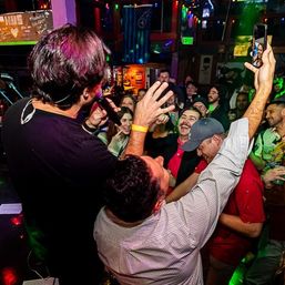 Crowded bar with a singer on stage engaging cheering fans as colorful party lights flash and a guest raises a phone to record a selfie-video.