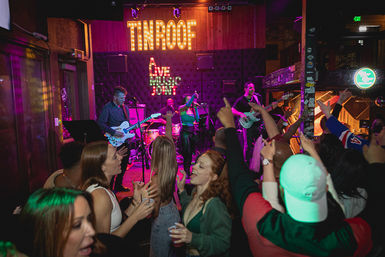 Vibrant nightlife scene: crowd cheering in a neon-lit indoor live music venue while a four-piece band performs on stage under marquee lights.