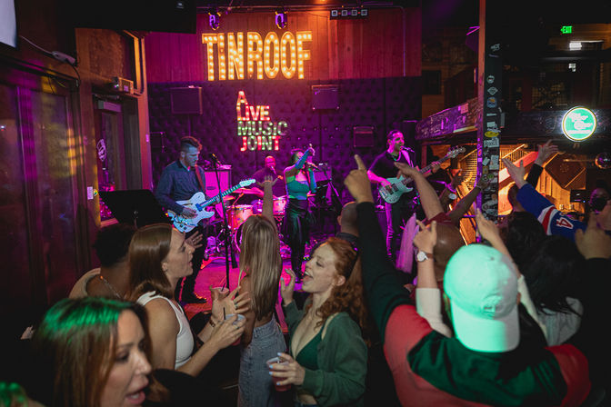 Vibrant nightlife scene: crowd cheering in a neon-lit indoor live music venue while a four-piece band performs on stage under marquee lights.