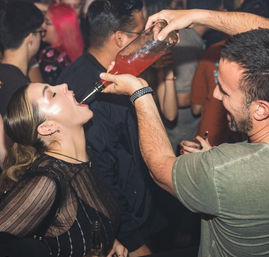 Lively nightclub scene with a man pouring a pink cocktail from a bottle into a woman’s open mouth amid a crowded bar.