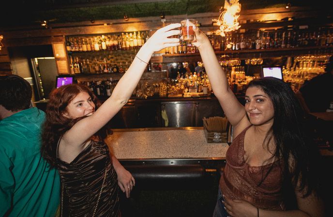 Two women toasting with shot glasses at a lively city bar — smiling friends enjoying late-night nightlife with warm lighting and rows of liquor bottles on the shelves behind the counter.