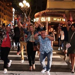 Group of friends cheering and dancing across a lit city crosswalk at night in a lively downtown nightlife district with street lamps and marquee lights in the background