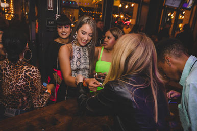 Group of young adults at a crowded nightclub bar having wristbands checked, laughing and holding drinks under warm string lights