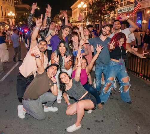 Cheerful group of young adults posing and waving on a lively downtown street at night, colorful bar and restaurant lights and festive nightlife atmosphere