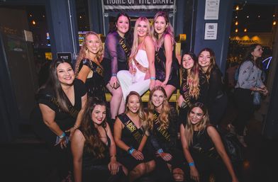 Cheerful bachelorette group of women in black dresses and sashes surrounding a bride-to-be in white, posing on an oversized yellow chair inside a lively downtown bar at night