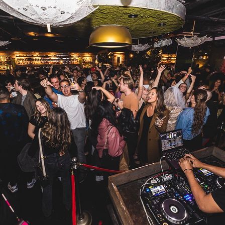 Packed nightclub dance floor with DJ booth and laptop in foreground, energetic crowd dancing and cheering under warm pendant lights and illuminated bar shelves