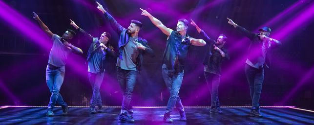 Six male dancers in casual streetwear strike a synchronized pointing pose on a theater stage bathed in purple spotlights