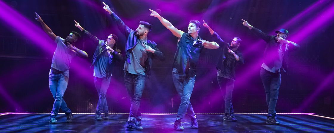 Six male dancers in casual streetwear strike a synchronized pointing pose on a theater stage bathed in purple spotlights