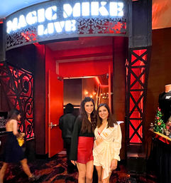 Two smiling women pose at a red-lit theater/nightlife entrance beneath a glowing marquee; one wears a celebratory sash, with holiday décor and a merch table visible nearby.