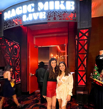 Two smiling women pose at a red-lit theater/nightlife entrance beneath a glowing marquee; one wears a celebratory sash, with holiday décor and a merch table visible nearby.