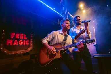 Live music duo on a smoky club stage: guitarist strumming an acoustic guitar while a vocalist sings into a microphone under blue and purple lights beside a red neon sign reading ‘FEEL MAGIC’.