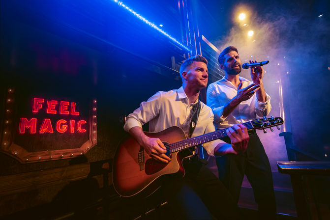 Live music duo on a smoky club stage: guitarist strumming an acoustic guitar while a vocalist sings into a microphone under blue and purple lights beside a red neon sign reading ‘FEEL MAGIC’.