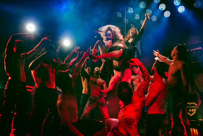 Nightclub stage show: a singer riding a plush unicorn prop as shirtless dancers lift and surround them under colorful spotlights in an energetic cabaret-style performance.