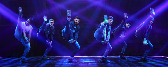 Six male dancers in urban outfits executing synchronized high kicks on a dark theater stage lit by vivid purple-blue spotlights and laser beams — energetic live choreography.
