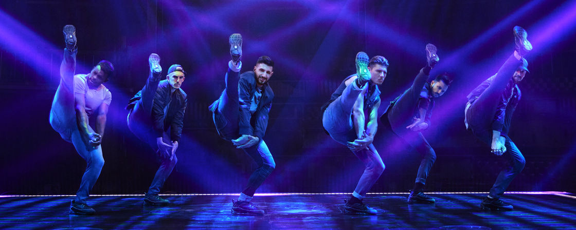 Six male dancers in urban outfits executing synchronized high kicks on a dark theater stage lit by vivid purple-blue spotlights and laser beams — energetic live choreography.