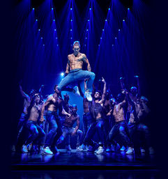 Shirtless male dancer in jeans and sneakers leaps above a shirtless ensemble on a theater stage under dramatic blue spotlights — high-energy contemporary dance performance.